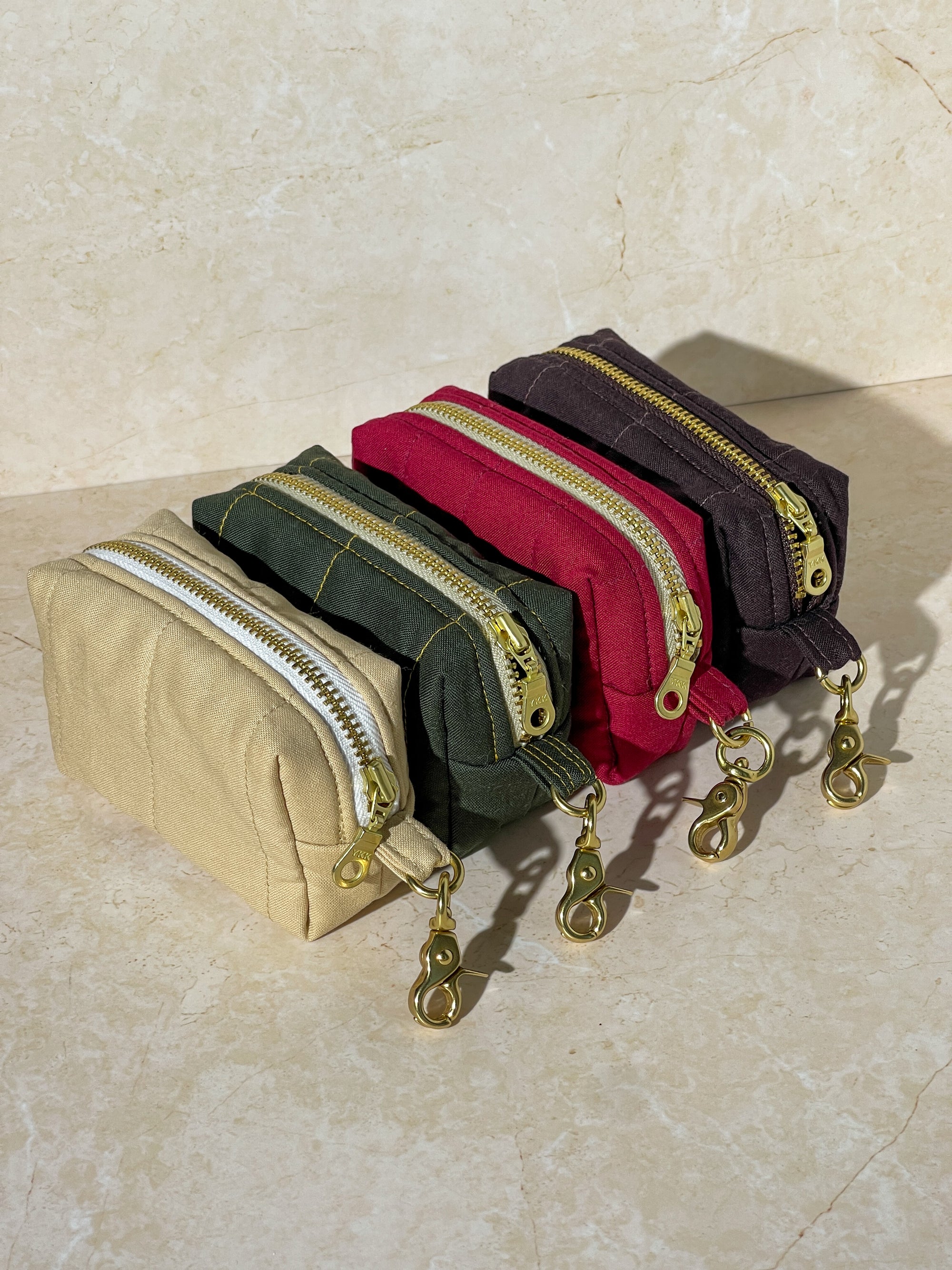 Four handmade fabric pouches with zippers in beige, green, red, and purple on a light stone background