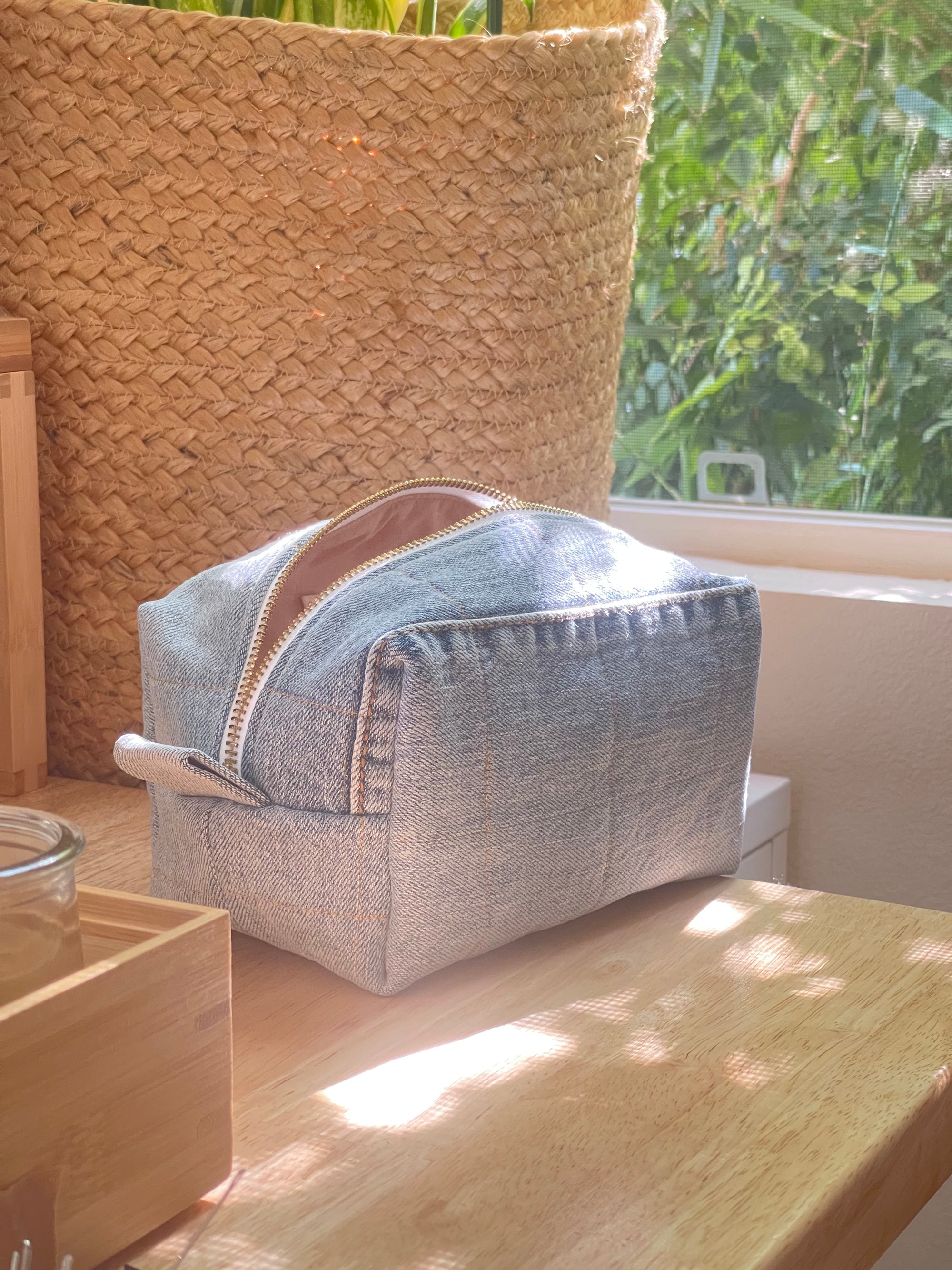 Upcycled light blue denim makeup bag on a wooden table next to a window with greenery outside