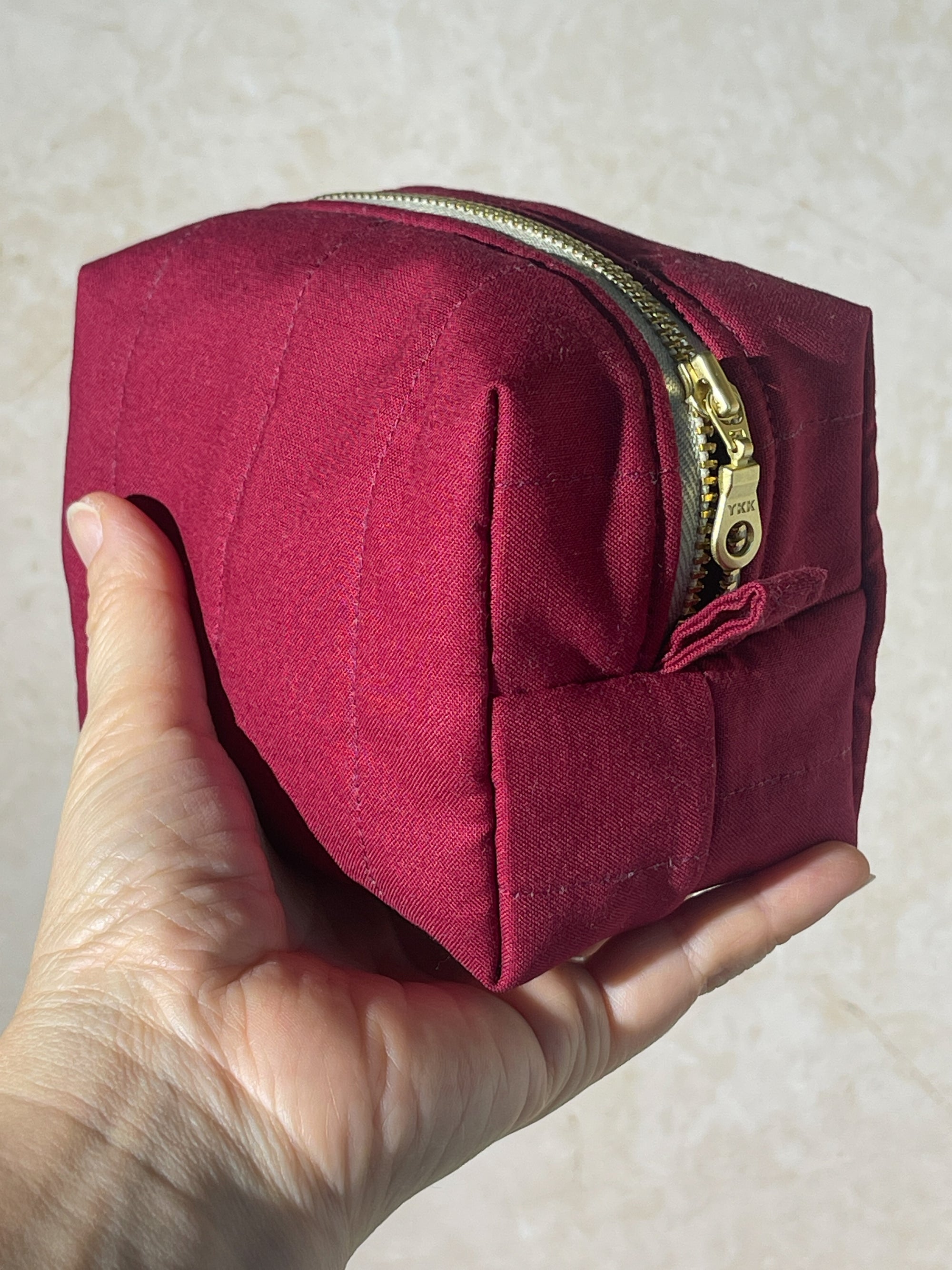 Hand holding a handmade small burgundy makeup pouch bag with gold zipper against a neutral background 