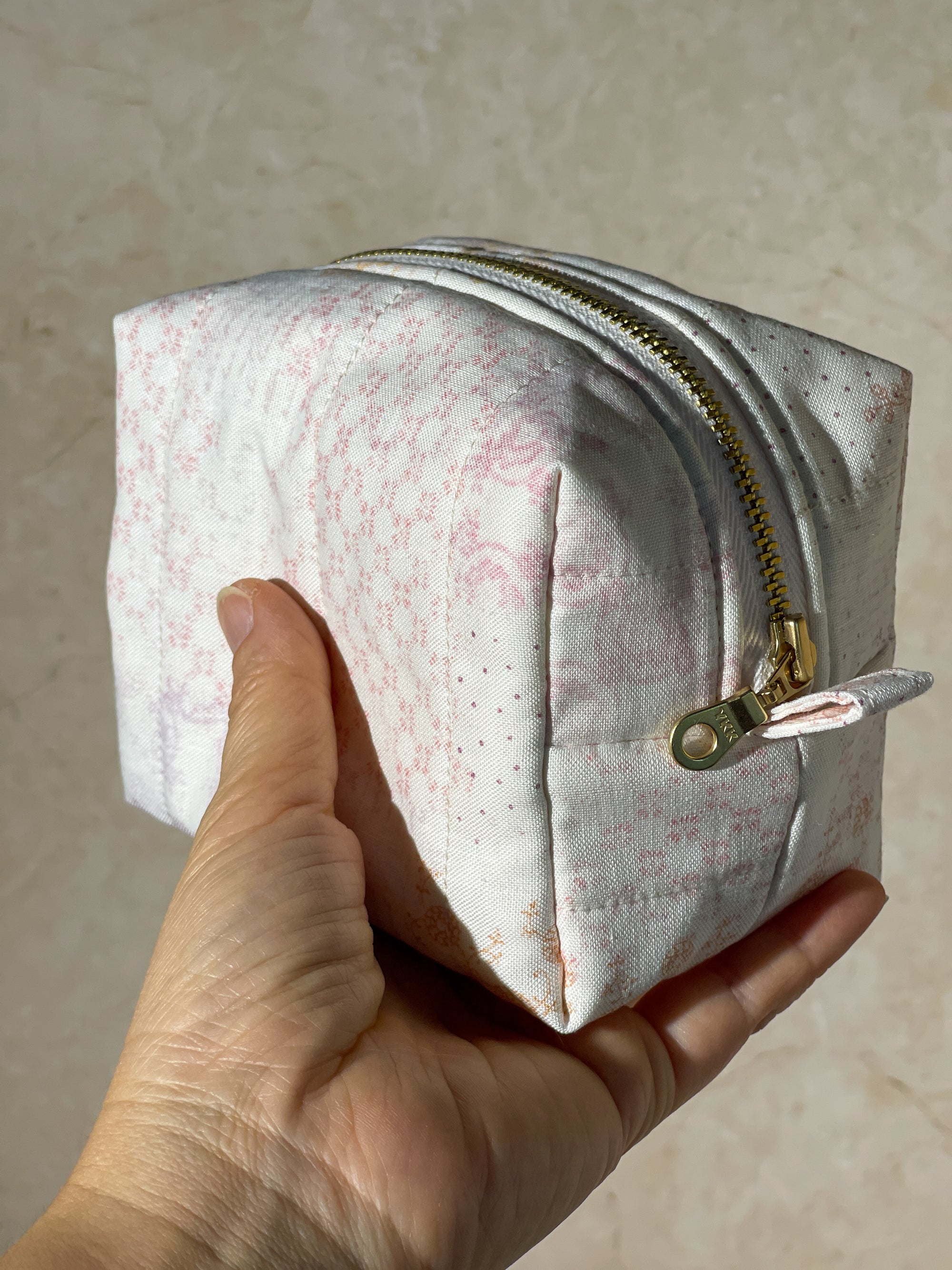 Hand holding a handmade small white makeup pouch with pink floral pattern and gold zipper against a beige background